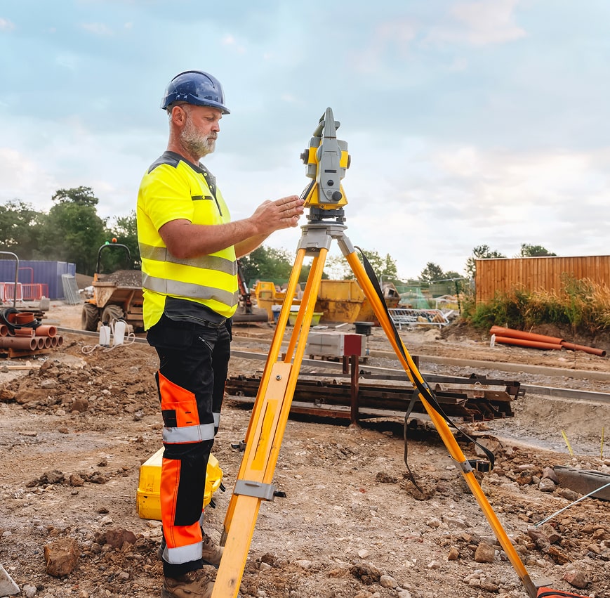 Man in hard hat and high-vis vest using surveying equipment on a construction site.
