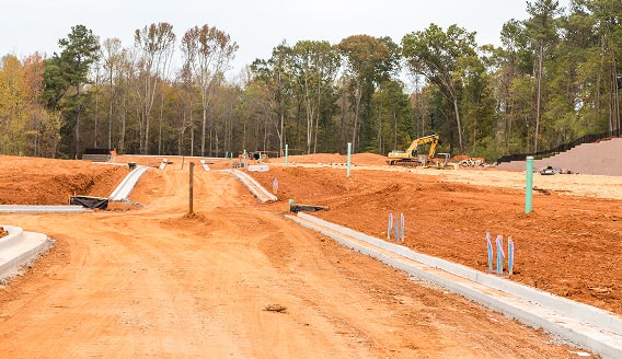 Construction site with dirt road and curbs, trees in background.